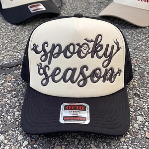 May include: A black and cream trucker hat featuring the embroidered text spooky season in a dark script font. The design includes small embroidered ghosts, a bat, a spider, and a spiderweb. A silver and black sticker on the brim reads OTTO.