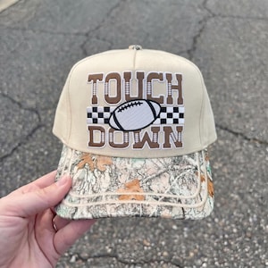 Op de afbeelding: Een beige honkbalpet met bruine geborduurde tekst TOUCH DOWN, een centrale football-afbeelding en een zwart-wit geblokt patroon. De klep heeft een gedetailleerde camouflageprint met bruine en beige bladeren.