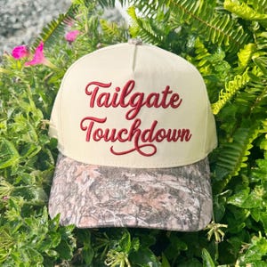 Może przedstawiać: Kremowa czapka z daszkiem z wyhaftowanym czerwonym napisem Tailgate Touchdown na przednim panelu. Czapka posiada usztywnianą koronę oraz zakrzywiony daszek z wyraźnym brązowo-szarym wzorem moro na całej powierzchni materiału.