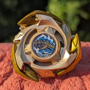 May include: A gold-coloured spinning top toy featuring a metallic finish and a central circular emblem. The emblem displays a stylised blue and gold dragon-like creature graphic with the letter X printed at the bottom of the design.