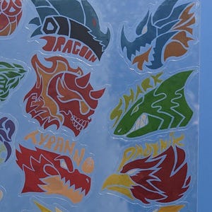 May include: A sheet of colorful stickers featuring various mythical creatures, including a dragon, a shark, a tyrannosaurus, and a phoenix. Each sticker has a unique design and includes the name of the creature in a stylized font.