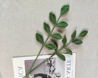 May include: Two green crocheted leafy stems with long brown stems. The stems are laying on a book with a photo of a person wearing a black sweater and jeans.