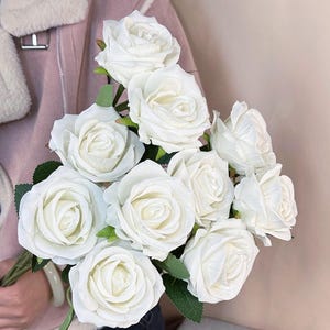 May include: A bouquet of white roses with green leaves. The roses are in full bloom, with multiple petals visible. The bouquet is held in front of a pink jacket.