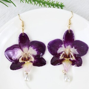 May include: A pair of handcrafted earrings featuring pressed purple orchid flowers with white and yellow accents. Each earring has a gold-colored hook and a dangling white pearl. The earrings are displayed on a white plate.