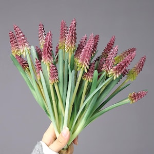 Puede incluir: Un ramo de flores artificiales con múltiples tallos y hojas largas y verdes. Las flores son de color burdeos intenso con un aspecto texturizado, que pasa a un verde más claro en las puntas. El ramo está atado con hilo de color natural.