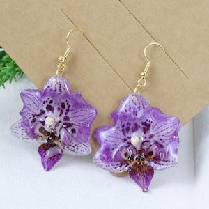 May include: A pair of dangle earrings featuring realistic purple orchid flower pendants with dark purple spots and a glossy finish. The earrings are attached to gold-toned metal fishhook wires and are displayed on a brown cardboard display card.