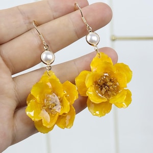 May include: A pair of dangle earrings featuring bright yellow flower charms with textured centers. Each earring is suspended from a gold-toned hook and accented with a single round white pearl bead positioned above the yellow floral element.