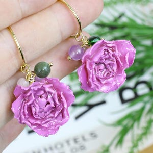 May include: A pair of gold hoop earrings featuring sculpted purple rose charms. Each earring is accented with small, round, multicolored beads in shades of green, light purple, and white, attached to the gold hoop near the rose flower.