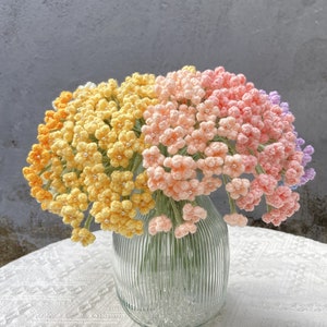 以下が含まれることがあります： 透明なガラスの花瓶に入った、手作りのかぎ針編みの花のブーケ。花は黄色、オレンジ、ピンク、紫のミックスで、各花の真ん中に小さな白いビーズが付いています。花瓶は白いレースのテーブルクロスの上に置かれています。