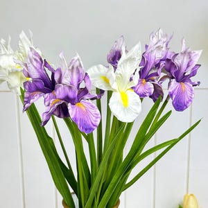 Puede incluir: Un ramo de flores de iris artificiales en un jarrón de cristal. Las flores son una mezcla de blanco y morado, con centros amarillos. El jarrón es gris oscuro con un borde dorado, y los tallos son verdes.