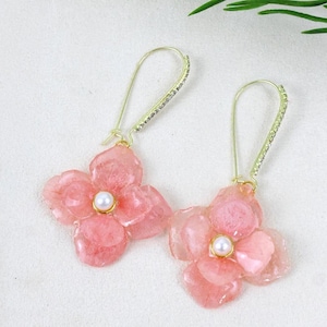 May include: A pair of gold-tone drop earrings featuring pink, translucent, flower-shaped resin charms with a central white pearl accent. The long, slender ear wires are embellished with a row of small, clear, sparkling crystals along the front.