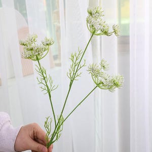 May include: A green and white artificial flower stem with multiple flower heads. The flower heads are small and clustered, with delicate white petals. The stem is held by a person in front of a white curtain.