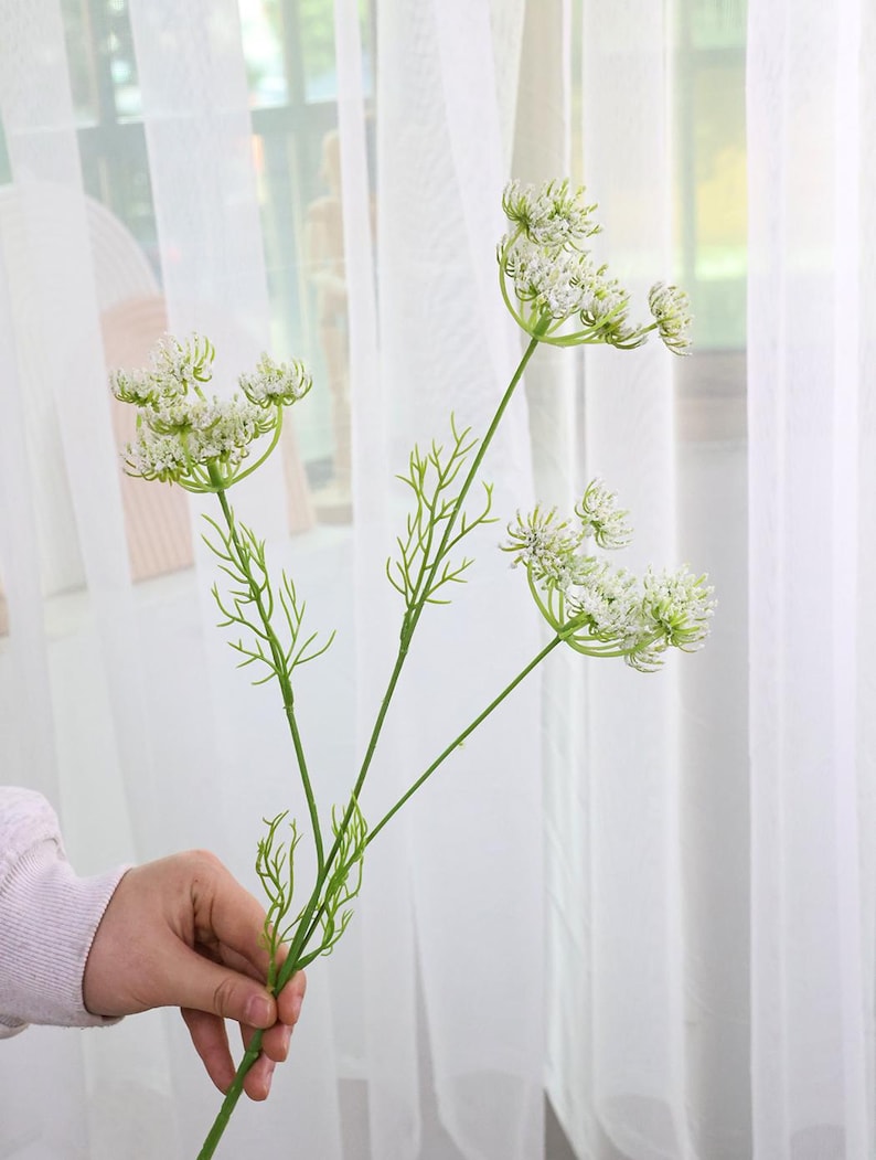 May include: A green and white artificial flower stem with multiple flower heads. The flower heads are small and clustered, with delicate white petals. The stem is held by a person in front of a white curtain.