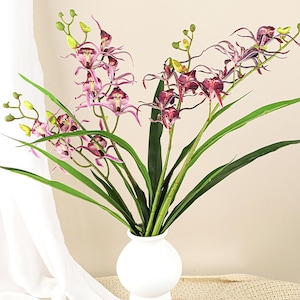 May include: A white vase displays a collection of artificial orchids. The flowers feature pink and burgundy petals, complemented by green stems and leaves. The vase has a minimalist design.