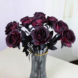 May include: A bouquet of dark red roses with black stems and leaves in a gray vase with a gold rim. The roses have a gradient color, transitioning from deep red to black. The arrangement is set against a neutral background.