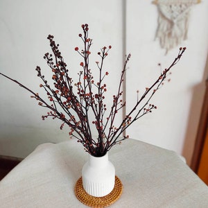 May include: Dried brown branches with small red berries arranged in a white vase with a ribbed design. The vase is sitting on a woven brown coaster.