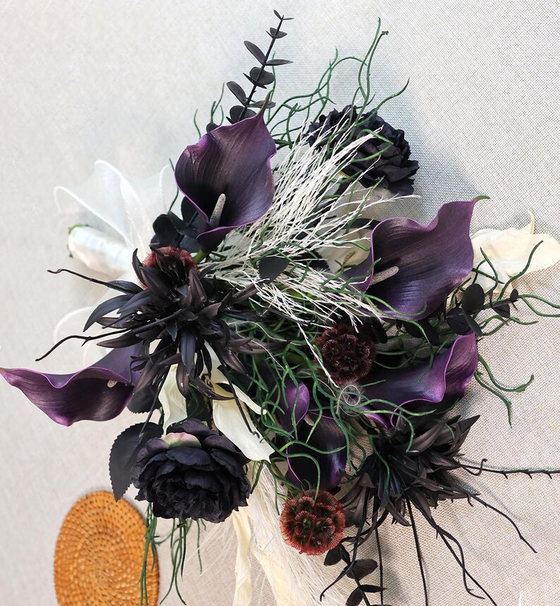 May include: A dramatic floral bouquet with deep purple calla lilies, black roses, and white accents. The arrangement incorporates various textures and shades of green, with a touch of white ribbon.