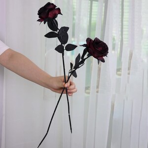 May include: Two artificial roses with deep burgundy blooms and black stems and leaves. The roses are held in a person's hand against a backdrop of a sheer white curtain and a window.