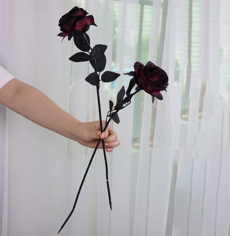 May include: Two artificial roses with deep burgundy blooms and black stems and leaves. The roses are held in a person's hand against a backdrop of a sheer white curtain and a window.