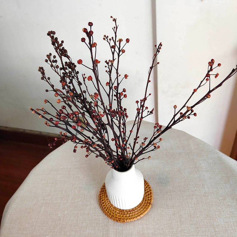 May include: Dried flower arrangement in a white vase with a woven coaster. The flowers are brown with small red berries.