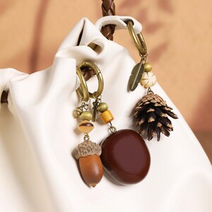 Puede incluir: Primer plano de dos pendientes de aro color bronce con abalorios con temática de la naturaleza. Un pendiente presenta una bellota, una piedra marrón y cuentas de madera. El otro tiene una piña, una pluma y cuentas color crema.