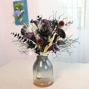 May include: A floral arrangement in a clear glass vase. The bouquet features dark purple calla lilies, black roses, and white accents. The vase is on a table with a light-coloured tablecloth.