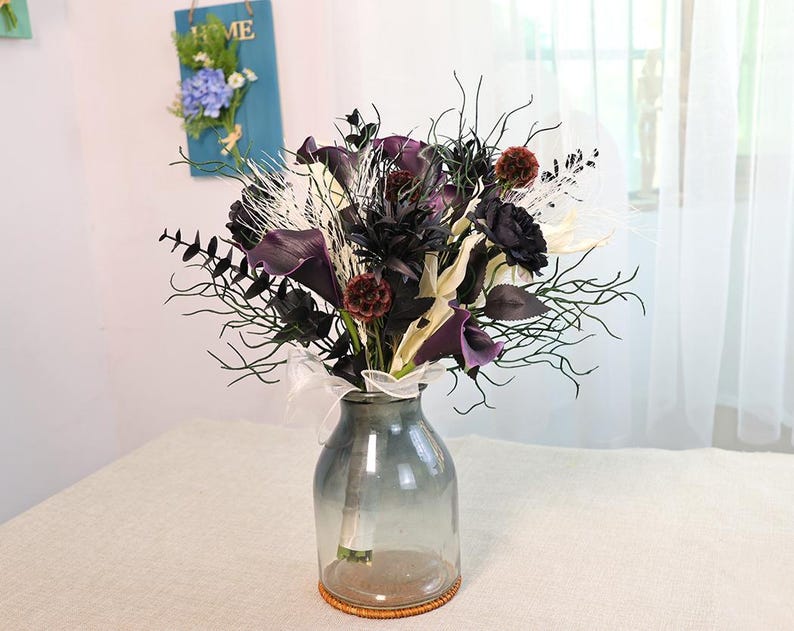 May include: A floral arrangement in a clear glass vase. The bouquet features dark purple calla lilies, black roses, and white accents. The vase is on a table with a light-coloured tablecloth.