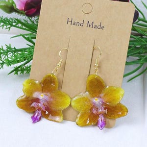 May include: A pair of dangle earrings featuring preserved yellow and brown orchid flowers with purple centers, attached to gold-toned metal hooks. The earrings are displayed on a rectangular brown card with the text Hand Made printed on it.