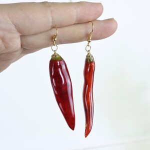 May include: A pair of dangle earrings featuring realistic red chili pepper charms. Each earring is suspended from a gold-toned metal hook. The chili peppers have a glossy, smooth finish with green stems at the top, showcasing distinct shapes.