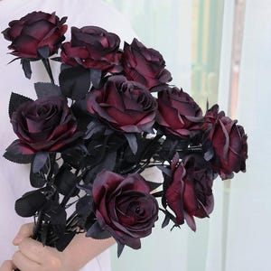 May include: A bouquet of dark burgundy artificial roses with black stems and leaves. The roses have multiple layers of petals, and the overall arrangement is held in a person's hand. The background is a soft, neutral color.