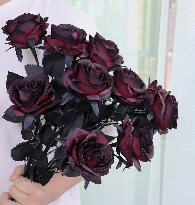 May include: A bouquet of dark burgundy artificial roses with black stems and leaves. The roses have multiple layers of petals, and the overall arrangement is held in a person's hand. The background is a soft, neutral color.