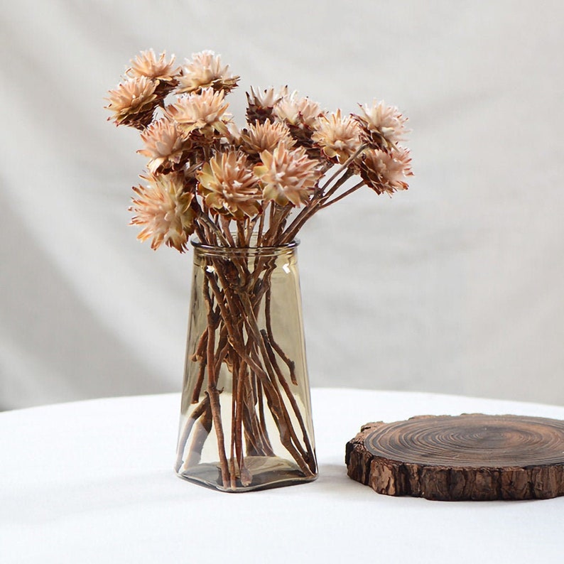 Op de afbeelding: Een boeket gedroogde bloemen in tinten van bruin en wit, gerangschikt in een heldere glazen vaas met een bruine tint. De vaas staat op een wit oppervlak met een houten schijf erachter.