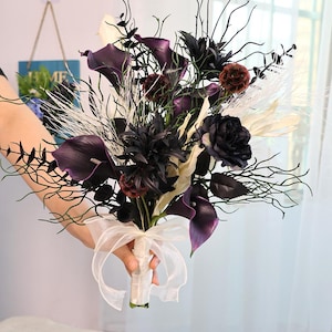 May include: A bouquet featuring dark purple calla lilies, black roses, and burgundy accents, tied with a cream-coloured ribbon. The arrangement includes black branches and white feathery elements, creating a dramatic floral display.