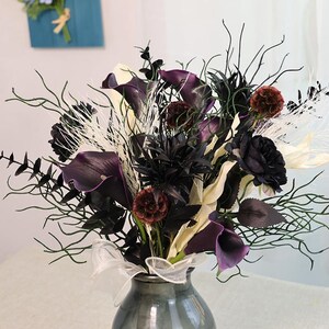 May include: A floral arrangement in a grey vase. The bouquet features dark purple calla lilies, black roses, and dried accents. White ribbon is tied around the vase. The arrangement includes dark green branches and white feathery elements.