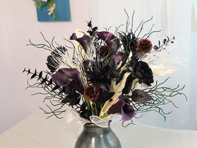 May include: A floral arrangement in a grey vase. The bouquet features dark purple calla lilies, black roses, and dried accents. White ribbon is tied around the vase. The arrangement includes dark green branches and white feathery elements.