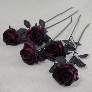 May include: Five artificial roses with deep burgundy petals and dark gray stems and leaves. The roses are in various stages of bloom, with some fully open and others partially closed. The stems are long and slender.
