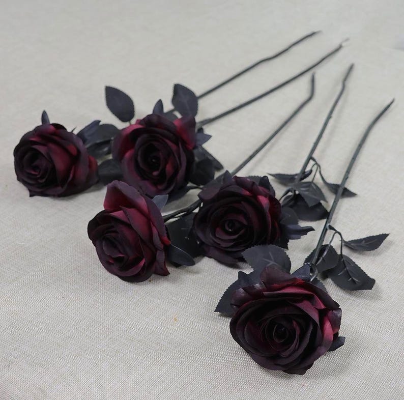 May include: Five artificial roses with deep burgundy petals and dark gray stems and leaves. The roses are in various stages of bloom, with some fully open and others partially closed. The stems are long and slender.