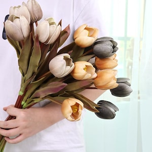 May include: A bouquet of artificial tulips in various colors, including cream, yellow, and dark gray. The faux flowers have green and burgundy leaves and stems. The bouquet is held by a person wearing a white shirt.
