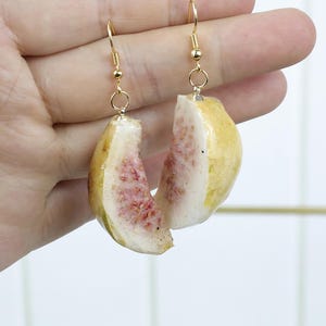 May include: A pair of dangle earrings featuring realistic, sliced guava fruit charms. Each charm displays a pale yellow rind and a textured, light pink interior with small seeds. The fruit pieces are attached to gold-toned metal fishhook wires.