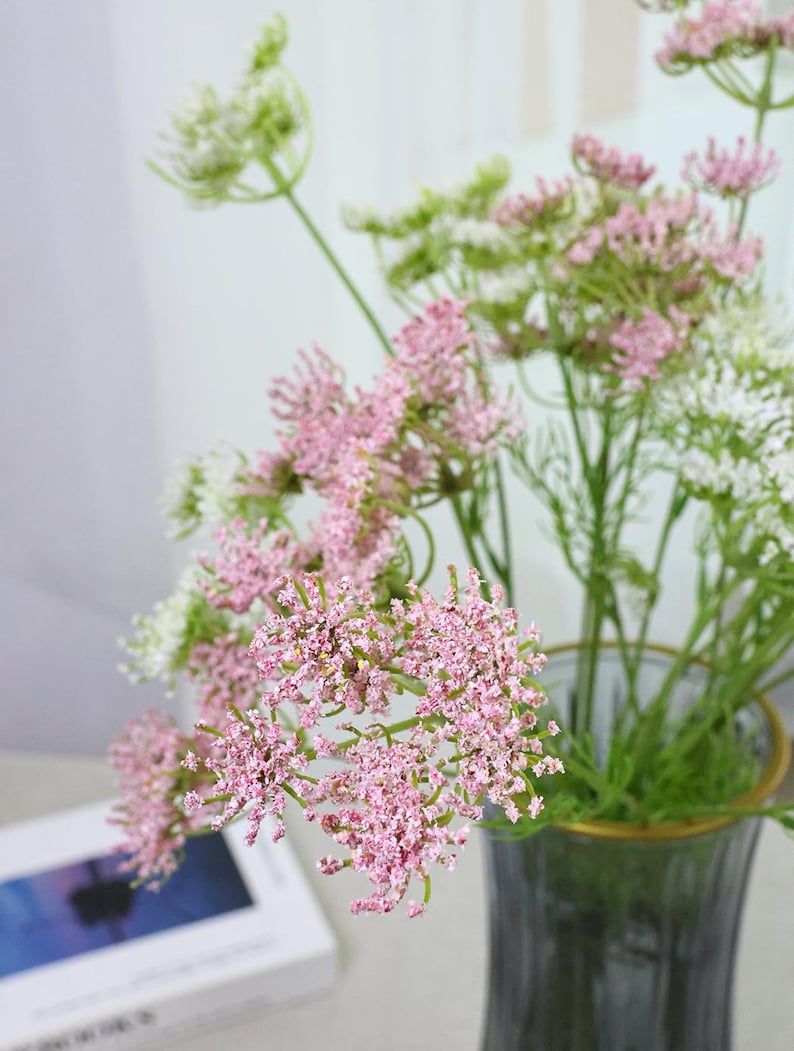 May include: A bouquet of pink and white Queen Anne's Lace flowers in a gray glass vase with a gold rim. The flowers have delicate, clustered blooms and green stems. A book is partially visible in the background.