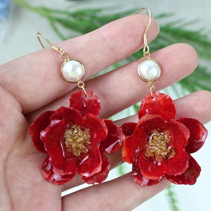 May include: A pair of handcrafted earrings featuring vibrant red flower pendants with gold-colored centers and gold-tone hooks. Each earring has a white pearl accent. The earrings are designed to be worn as a fashion accessory.