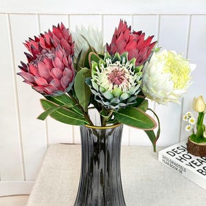 Könnte beinhalten: Ein Strauß künstlicher Protea-Blumen in einer grauen Glasvase. Die Blumen sind in verschiedenen Farben gehalten, darunter Rot, Weiß und Grün. Die Vase steht auf einer beigen Oberfläche. Ein Buch mit dem Text "DESIGN HOTELS YEAR BOOK 08" ist sichtbar.