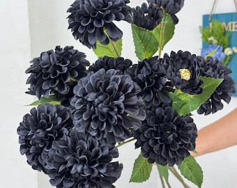 May include: A bouquet of dark gray artificial dahlia flowers with green leaves. The flowers have multiple layers of petals, and the stems are visible. The bouquet is held against a white background.