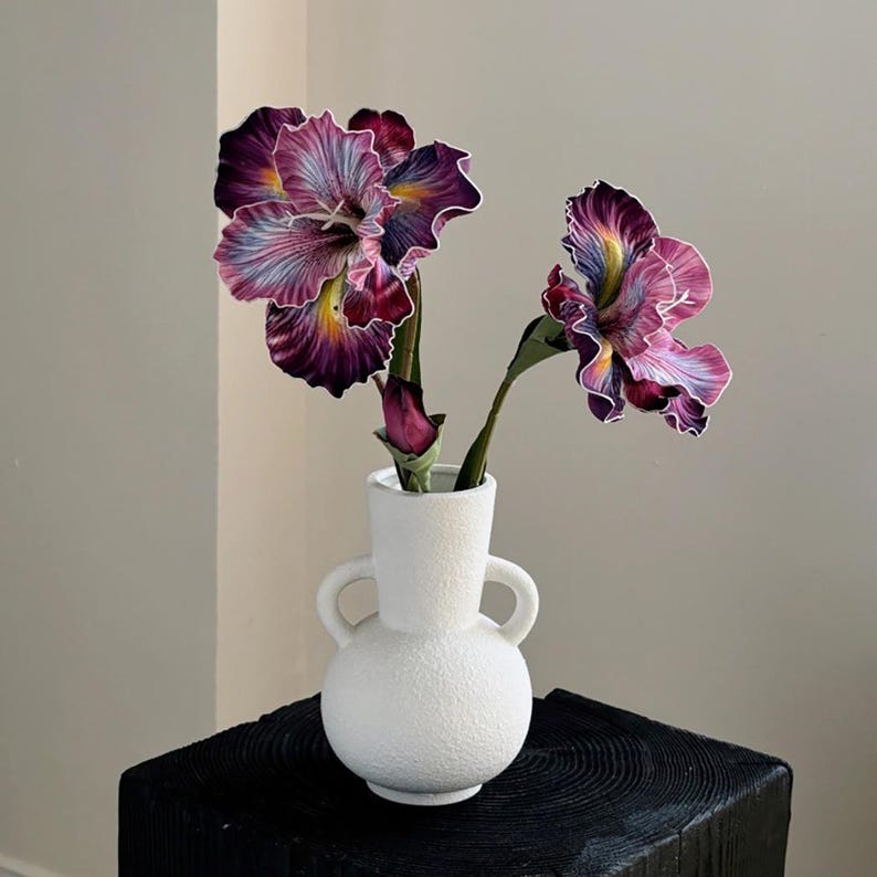 May include: Two purple and white iris flowers in a white ceramic vase with two handles. The vase has a textured surface.