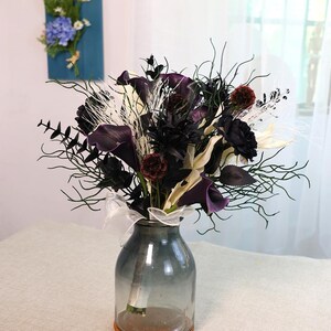 May include: A bouquet of dark purple calla lilies, black roses, and other dark-coloured flowers and foliage in a clear glass vase. The vase sits on a woven coaster and is tied with a white ribbon.