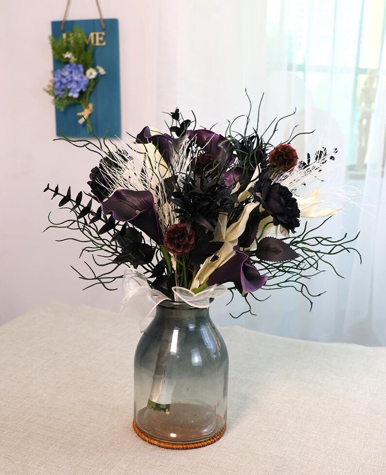 May include: A bouquet of dark purple calla lilies, black roses, and other dark-coloured flowers and foliage in a clear glass vase. The vase sits on a woven coaster and is tied with a white ribbon.