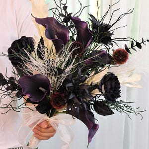 May include: A dramatic bouquet with deep purple calla lilies, black roses, and cream-coloured leaves. The arrangement includes white sprigs and is tied with a cream ribbon. A striking floral display, ideal for a wedding or special occasion.