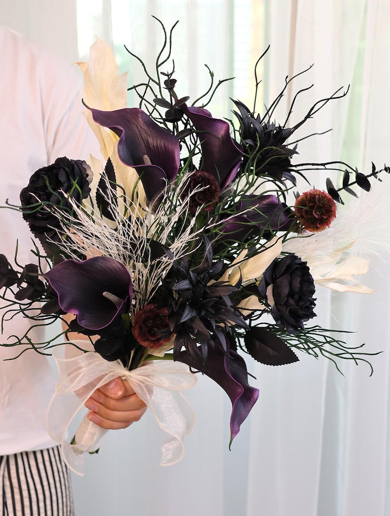 May include: A dramatic bouquet with deep purple calla lilies, black roses, and cream-coloured leaves. The arrangement includes white sprigs and is tied with a cream ribbon. A striking floral display, ideal for a wedding or special occasion.