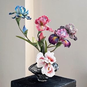 Peut inclure: Un bouquet de cinq fleurs d'iris en soie dans un vase en verre transparent avec une surface texturée. Les fleurs sont dans des tons de bleu, rose, violet et blanc.