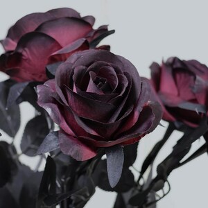 May include: Close-up of artificial roses with deep burgundy petals and dark gray leaves. The roses have a velvety texture and are in full bloom, showcasing their intricate layers. The stems are a dark gray color, and the background is a soft white.